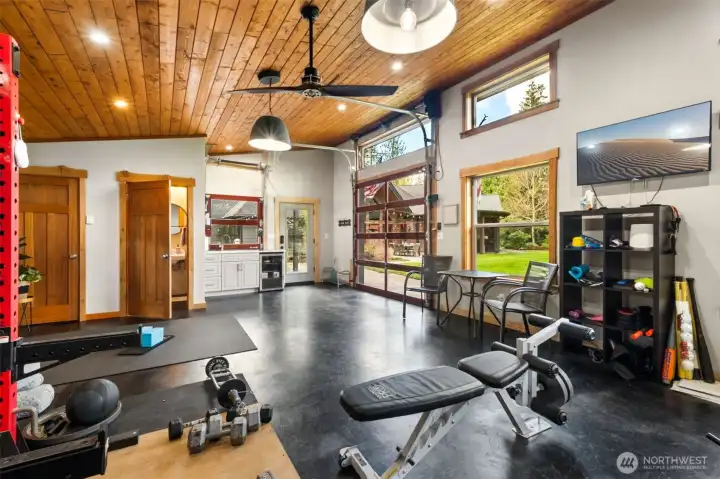 Currently used as an exercise space, the cabana’s interior showcases vaulted tongue-and-groove wood ceilings, contemporary lighting, and ceiling fans for comfort. Clerestory windows bring in additional natural light, while the glass garage door opens wide to the outdoors—perfect for gatherings, workouts, or studio use.