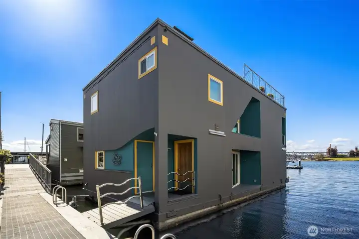 The front façade from the dock: modern, gray-on-teal, with mustard-trimmed window reveals and a glimpse of the rooftop deck overhead.