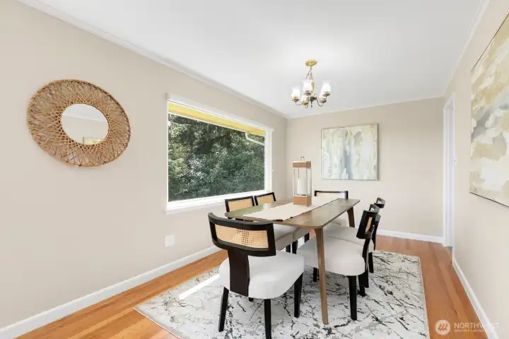 Sunlit dining room with chandelier, hardwood floors, and a large picture window framing lush greenery.