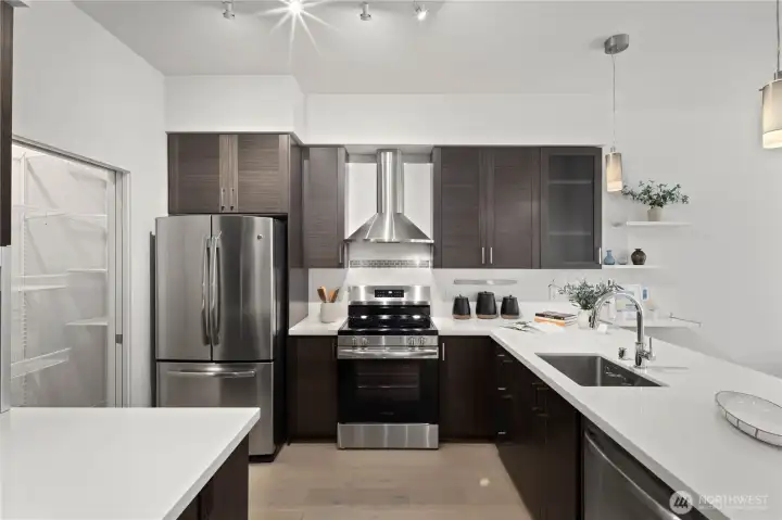 Chef-inspired kitchen featuring slab quartz-stone countertops with a waterfall edge, stainless steel appliances, a contemporary exhaust hood, custom wood cabinetry, and a spacious pantry.