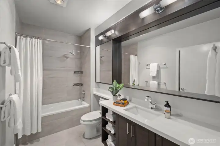Full bathroom featuring slab quartz-stone countertops, an undermount sink with a designer faucet, warm wood cabinetry, a custom mirrored storage cabinet, and a discreet stackable front-load washer and dryer.