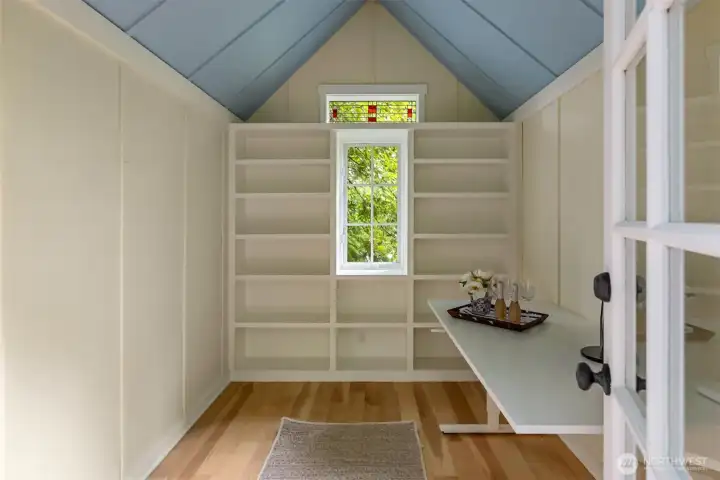The inside of this darling writer's studio with built-in shelves and a stained glass window above.