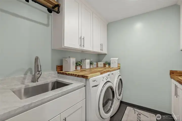 Spacious laundry room with built in cabinet storage, butcher block countertops, large utility sink, and bar to hang clothes.Tile in Laundry Room. The laundry room is equipped with a Whirlpool Duet HT(High-Efficiency/Thermal) front-loading washer and dryer.