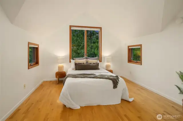 With its vaulted ceiling and symmetrical layout, the primary bedroom offers a sense of balance, calm, and quiet retreat. Three thoughtfully placed windows bring in lush green views from multiple angles, creating a treehouse-like feel. The natural hardwood flooring adds warmth, while the simple lines and airy proportions make the room feel open yet cozy. It’s a space designed for rest, reflection, and waking up grounded in nature.