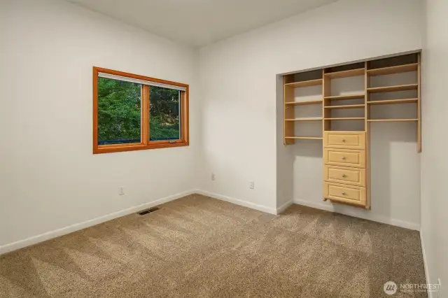 Guest Bedroom 1 on the main floor offers a peaceful, tree-framed view and a comfortable, versatile layout. Soft carpeting underfoot and neutral tones create a cozy atmosphere, while the built-in closet system provides practical storage with drawers and adjustable shelving. Whether used as a guest room, home office, or hobby space, this room benefits from natural light and easy access to the rest of the main living areas.