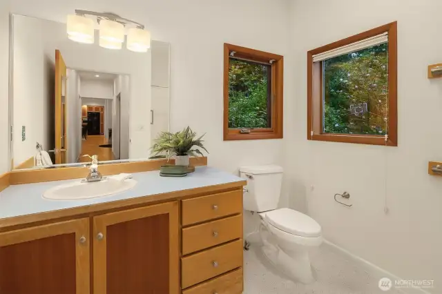 The main floor guest bath is clean, bright, and well-appointed with thoughtful touches throughout. Natural light pours in through two windows, offering a peaceful view of the surrounding greenery while maintaining privacy. The vanity provides generous counter space and storage, and updated lighting adds a warm, modern touch. Conveniently located near both guest bedrooms and the main living areas, this bathroom is perfectly positioned for visitors or everyday use.