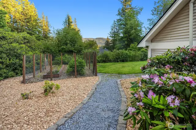 A gravel path leads gracefully from the driveway to the backyard garden area, bordered by flowering shrubs and natural landscaping. The fully fenced garden beds are ready for your vegetables, herbs, or cut flowers—protected and well-positioned to soak up the sun. Beyond the garden, a spacious lawn opens to mountain views and mature trees, creating a peaceful, usable outdoor space that invites both productivity and relaxation.