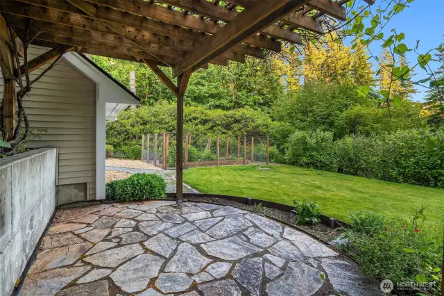 Tucked just off the lawn, this charming flagstone patio sits beneath a rustic grape arbor, offering a shaded spot to relax, dine, or garden in peace. The lush surroundings and gentle curve of the landscaping create a natural sense of enclosure and privacy. To the left, a dedicated garden shed blends seamlessly into the home’s exterior—perfect for storing tools, equipment, and seasonal supplies without cluttering the main living spaces. It’s a functional and serene backyard retreat designed for both beauty and utility.