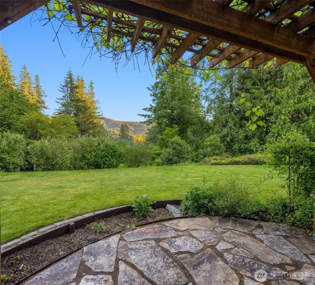 From beneath the vine-covered arbor, this view captures the expansive lawn and forested backdrop that define the home’s peaceful setting. The flagstone patio invites morning coffee or evening relaxation while overlooking mountain ridges in the distance. Framed by mature landscaping and surrounded by nature, the space feels like a private garden retreat—ideal for quiet moments, outdoor gatherings, or simply enjoying the beauty of the Pacific Northwest.