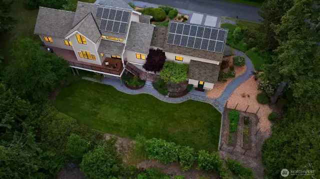 This aerial perspective showcases the home’s exceptional layout and the beauty of its landscaped surroundings. The expansive backyard features a lush, level lawn bordered by natural greenery and gardens, with stone and gravel pathways connecting the home to the fenced raised beds and grape arbor patio. Solar panels across the roof reflect a commitment to energy efficiency, while the thoughtful design of the outdoor spaces offers room to relax, grow, and entertain. Nestled into its half-acre+ lot, the property feels both private and purposeful—an ideal blend of modern living and Pacific Northwest charm.