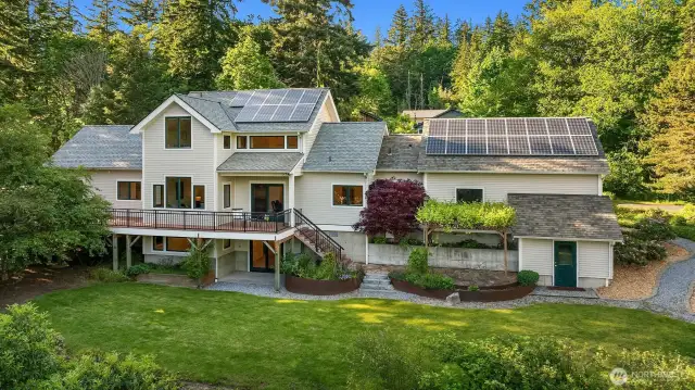 The back of the home reveals its full connection to the landscape, with a wide lawn, forested backdrop, and thoughtful outdoor living spaces. A large elevated deck extends from the main floor, offering sweeping views and seamless indoor-outdoor flow—perfect for entertaining or quiet evenings above the trees. Below, curved garden beds and stone paths create structure and beauty, while solar panels above speak to the home's energy-conscious design. With mature trees, privacy, and room to roam, the backyard is both functional and restorative.