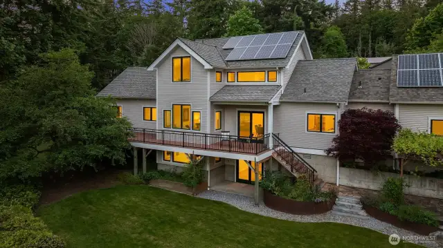 As evening sets in, the back of the home glows warmly through its many windows, highlighting the inviting atmosphere within. The elevated deck becomes a perfect perch to enjoy sunset views or stargazing, while landscape lighting softly illuminates the surrounding gardens and pathways. This twilight view captures the home’s peaceful setting and thoughtful design—blending comfort, sustainability, and a strong connection to the outdoors. It’s a place that feels just as welcoming at dusk as it does in the daylight.