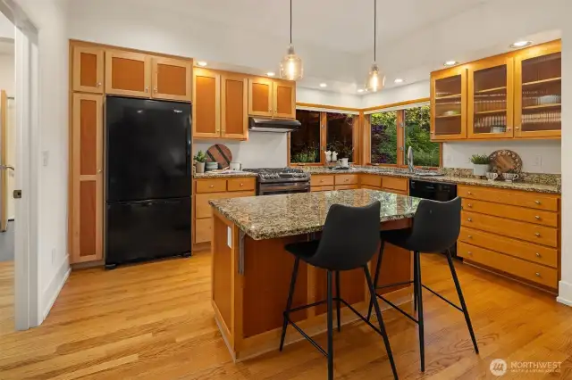 This kitchen combines warm natural wood cabinetry with sleek granite countertops and a functional layout that’s ideal for everyday use. The large corner windows bring in ample natural light and offer a pleasant view while at the sink. A center island with bar seating adds workspace and casual dining options. The black appliance package—including a gas range and French-door refrigerator—contrasts nicely with the wood tones for a clean, cohesive look. With abundant storage, under-cabinet lighting, and hardwood flooring throughout, this space is both practical and inviting.