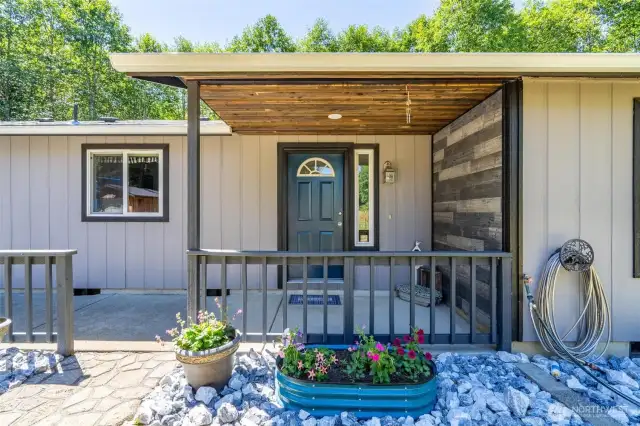 Welcoming front entry with charming curb appeal, featuring a deep blue front door, wood-accented ceiling, and stylish horizontal plank detailing. The covered porch offers protection from the elements while creating a cozy place to greet guests or enjoy a quiet moment outdoors. Thoughtful landscaping with stone accents and colorful planters adds warmth and personality. Whether you're arriving home or welcoming visitors, this inviting entry sets the tone for comfort, care, and country living.