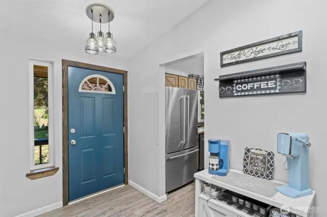 This light-filled foyer offers direct sightlines into the heart of the home, anchored by a sleek stainless steel French door refrigerator and custom wood cabinetry just beyond. A designated coffee and beverage station provides the perfect space-saving solution for morning rituals or hosting. The bold contrast of the navy front door and dark-stained trim frames the entry beautifully, while the triple pendant fixture adds a modern industrial flair. Functional meets cozy in this creative use of space, where everyday convenience gets a stylish upgrade.