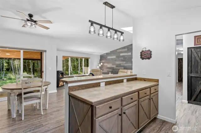 Seamless open-concept living with clear sightlines from the kitchen to the dining and living areas—ideal for entertaining, family gatherings, or staying connected while cooking. The extended breakfast bar with dual-height counters and ample storage doubles as a prep space or buffet station. Enjoy luxury vinyl plank flooring throughout, a cozy wood stove with stacked log storage, and a feature wall with rustic wood-look paneling that adds warmth and texture. Sliding glass doors lead to the covered patio, creating effortless indoor-outdoor flow in this thoughtfully designed home.