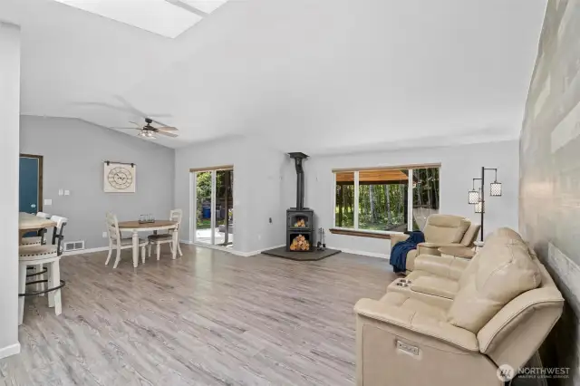 Spacious living room with vaulted ceilings, wood-burning stove, and panoramic forest views through oversized windows. This light-filled space is perfect for cozy nights by the fire or relaxing with a book while soaking in the serene scenery. The modern wood-look accent wall adds warmth and texture, while luxury vinyl plank flooring ensures durability and style throughout. Sliding glass doors extend the living space outdoors, creating the ideal blend of comfort, nature, and everyday livability.