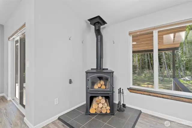 Efficient wood-burning stove offers both rustic charm and practical warmth, creating a cozy focal point for the main living area. Set on a tiled hearth and paired with built-in log storage and fireplace tools, this setup is ideal for chilly Pacific Northwest evenings. Large windows and nearby sliding glass doors frame peaceful wooded views, blending comfort and nature in one inviting corner of the home. Whether heating the space or adding ambiance, this feature enhances both sustainability and style.