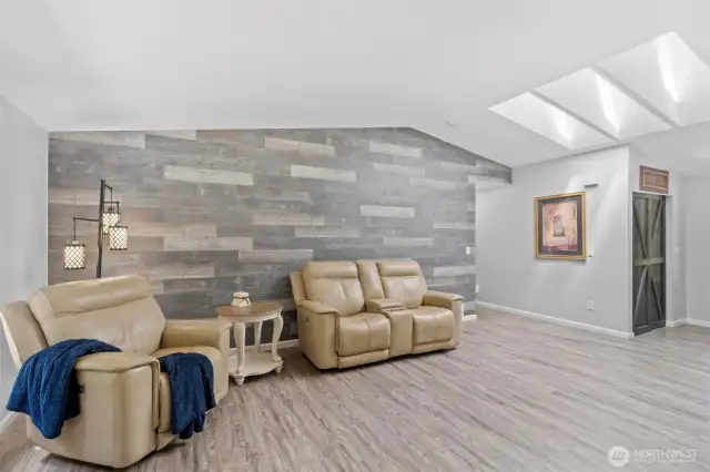 Modern farmhouse living room with stunning wood-look accent wall, vaulted ceiling, and triple skylights that flood the space with natural light. The wide plank luxury vinyl flooring adds a cohesive and durable foundation, while neutral tones provide a serene backdrop for your personal style. This cozy corner is ideal for relaxing, watching a movie, or reading by soft natural light. With open sightlines to the kitchen and entry, this space offers both comfort and continuity throughout the home.