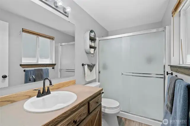 Attached 3/4 en suite bathroom features a large step-in shower with frosted glass sliding doors, stylish matte black fixtures, and a wood-accented vanity with ample storage. Natural light filters in through the window, enhancing the bright and clean aesthetic. Durable finishes and thoughtful design make this private bath both practical and inviting—perfect for everyday comfort and convenience within the primary suite.