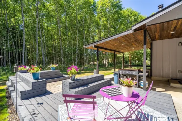 This thoughtfully designed outdoor space offers the ultimate blend of functionality and charm, with a covered patio featuring a rich wood-paneled ceiling and a connected open-air deck for seamless indoor-outdoor living. Built-in benches provide extra seating and visual interest, while a cozy bistro table invites alfresco dining or morning coffee in the sun. Surrounded by tall Pacific Northwest trees, this backyard retreat delivers unmatched privacy and a true connection to nature. Ideal for entertaining, grilling, or unwinding in your own quiet woodland sanctuary.