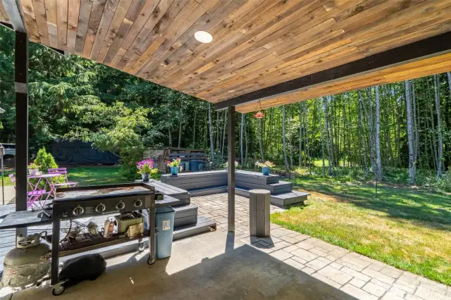 Covered patio with beautifully finished wood ceiling offers the perfect shaded zone for outdoor cooking, relaxing, or entertaining guests. Enjoy grilling, dining, or lounging while taking in peaceful views of the tree-lined yard and lush natural surroundings. Built-in bench seating adds functionality and charm to the extended deck area, while the tranquil backdrop of Pacific Northwest forest creates a true sense of privacy. Whether hosting weekend BBQs or enjoying quiet mornings with birdsong and coffee, this backyard sanctuary offers a lifestyle rooted in nature and comfort.