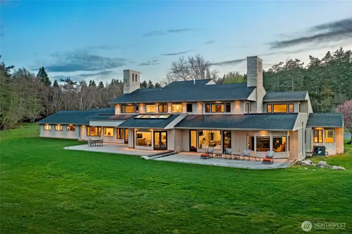 Twilight view of the estate showcasing warm interior glow, dual stone chimneys, and expansive patio spaces designed for elegant entertaining on San Juan Island.