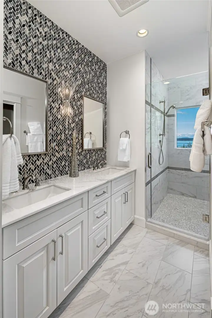 Elegant bath with dual vanity, marble countertops, designer tile accent wall, and glass-enclosed walk-in shower with island and water views.