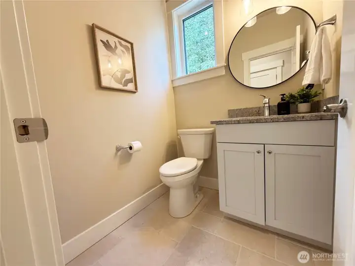 POWDER ROOM ON MAIN. This powder room presents a bright, clean, and thoughtfully designed space with a warm, welcoming feel. Soft neutral walls are paired with light tile flooring, creating an airy and cohesive look. A white vanity with simple, classic cabinetry is topped with a stone countertop, complemented by a round mirror and modern lighting that adds a subtle contemporary touch.