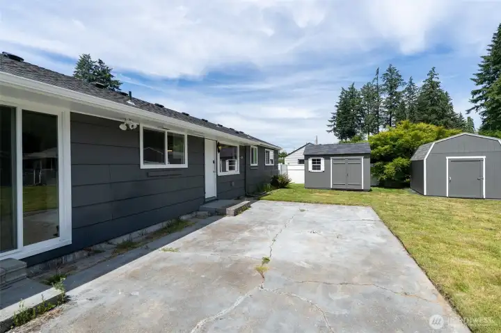 View of back of home showing huge patio, patio doors leading out from your rec room, and two of your outbuildings. Yard is fully-fenced and larger than most!