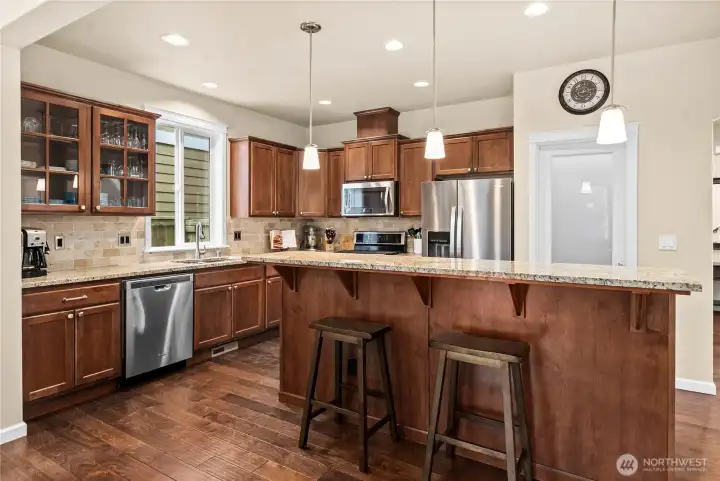 Spacious kitchen with granite countertops, rich cabinetry, stainless steel appliances, and a large island with seating and abundant storage on the opposite side.