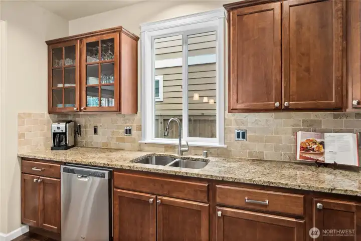 Large window above the stainless steel double sink fills the kitchen with natural light, complemented by full tile backsplash and elegant glass-front cabinetry.