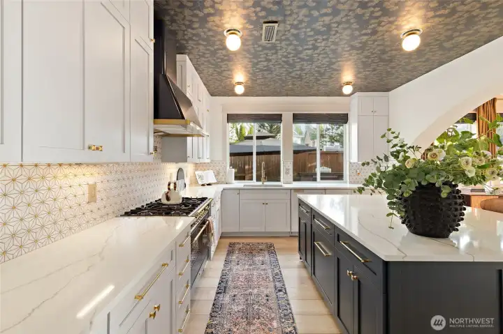 As you continue into the kitchen, the thoughtful design details carry through—note the abundance of prep space, natural light pouring in from the oversized windows, and the striking contrast of the dark island against the light custom cabinetry. The patterned ceiling adds unexpected drama, tying together the luxe finishes showcased throughout this beautifully curated home.