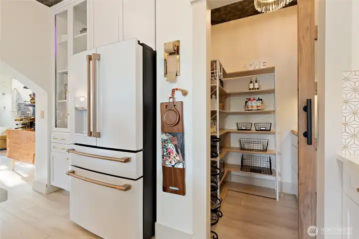 Just off the kitchen, the walk-in pantry offers both beauty and function. Thoughtfully designed with custom-stained wood shelving, a butcher block countertop with integrated outlets, and ample cabinetry, it’s as practical as it is polished. The space is elevated with a bold ceiling wallpaper and a sparkling brushed brass crystal flush mount light fixture. A frosted glass barn door adds just the right touch of modern elegance while keeping everything tucked away yet easily accessible.