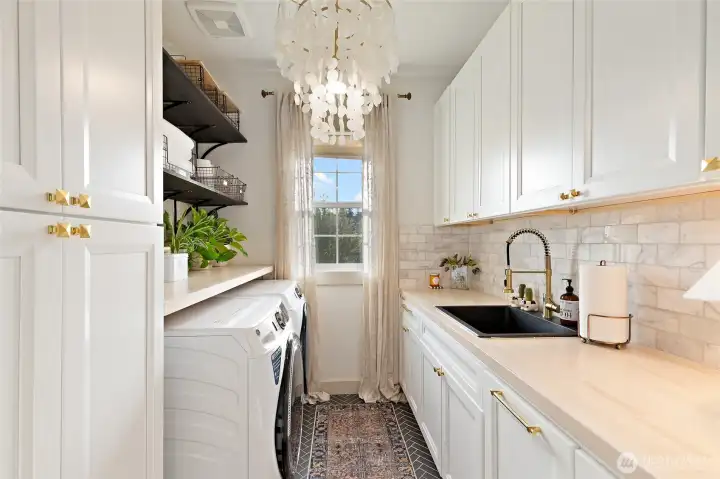 Laundry day just got a serious upgrade. This thoughtfully redesigned space features a 4.8 cu. ft. front-load washer with UltraFresh Vent System and Sanitize with Oxi, set beneath an extended butcher block counter for easy folding and organization. Soft-close white cabinetry, brushed gold hardware from Rejuvenation, and a marble tile backsplash elevate the entire room, while a built-in sink adds function to the form. Herringbone tile flooring and a timeless chandelier bring unexpected elegance, and open wood-stained shelving keeps essentials both accessible and stylish. A rustic sliding barn door completes the space, proving that utility can be just as beautiful as any other room in the home.