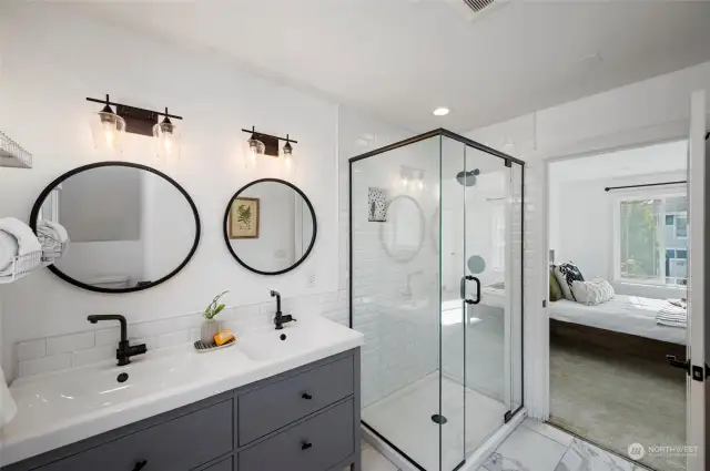 En suite bathroom with a walk-in tile shower, dual sinks, sleek black fixtures, and tile floors.