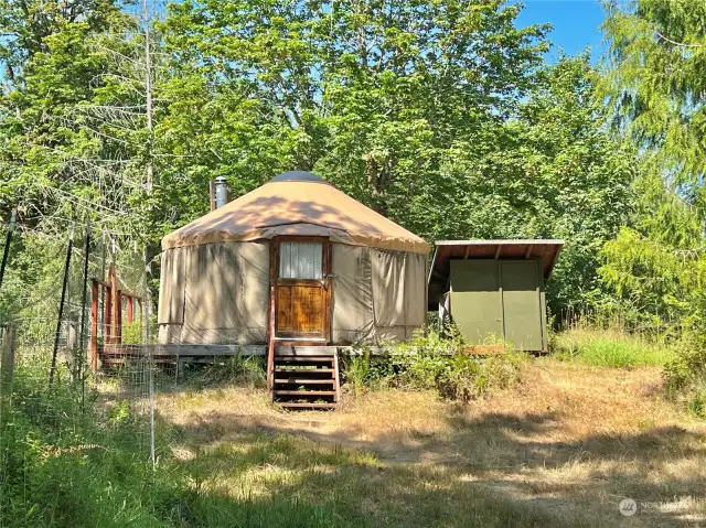 Front of Yurt. Notice the Wood Shed to the right, the wooden deck that encompasses the yurt, and the fenced garden to the left with apple and plum trees.