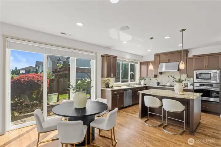 Bright, open kitchen and dining space with large windows, sliding glass doors, and backyard views. A granite island with seating, rich wood cabinetry, and abundant natural light creates an inviting everyday gathering space.