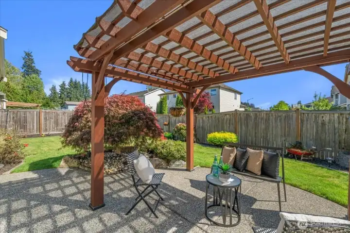 Inviting covered patio with pergola-style overhead structure, creating an ideal outdoor space for dining, relaxing, or entertaining. Overlooks the landscaped backyard with mature plantings and lawn area.
