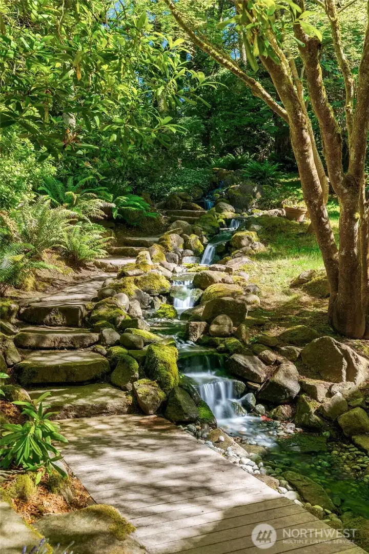 A beautifully crafted cascading water feature winds through lush gardens and beneath a charming footbridge, creating an unforgettable approach to the home and a soothing soundtrack to everyday life