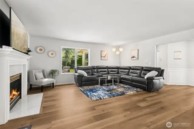 Cozy Family Room with Fireplace - spacious family room featuring a modern gas fireplace with marble surround, luxury vinyl plank flooring, and large picture windows that bring in natural light and garden views. Ideal for relaxing or hosting gatherings in comfort.