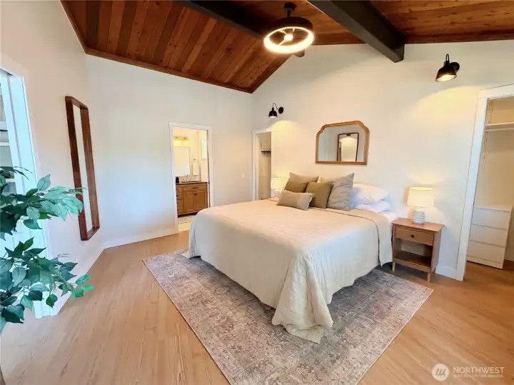 Another view of the serene primary suite showcases the vaulted wood ceiling, private ensuite bath access, walk-in closet, and the warm, peaceful atmosphere that makes this retreat so special. Deck access with lake views is just steps away.