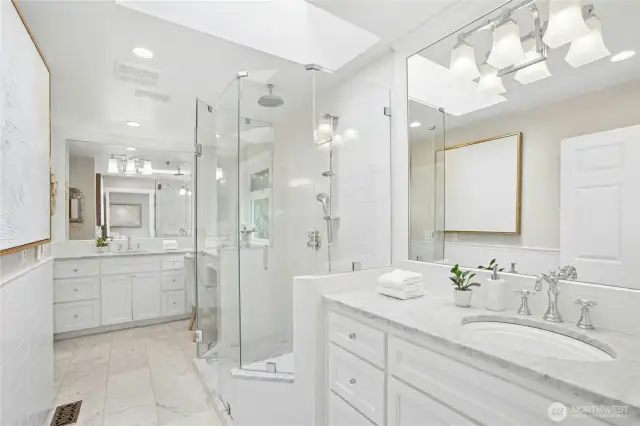 Spa-inspired retreat featuring heated floor, dual marble-topped vanities, frameless glass shower, and designer lighting beneath a skylit ceiling.