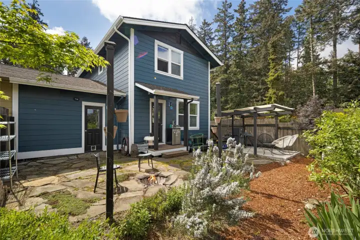This private backyard oasis features a stone patio with a cozy fire pit, mature landscaping with blooming rosemary and lush greenery, and a pergola-covered seating area — all framed by the home's bold blue siding, crisp white trim, and a backdrop of towering Pacific Northwest conifers. The covered back entry with direct patio access makes outdoor entertaining effortless and inviting.
