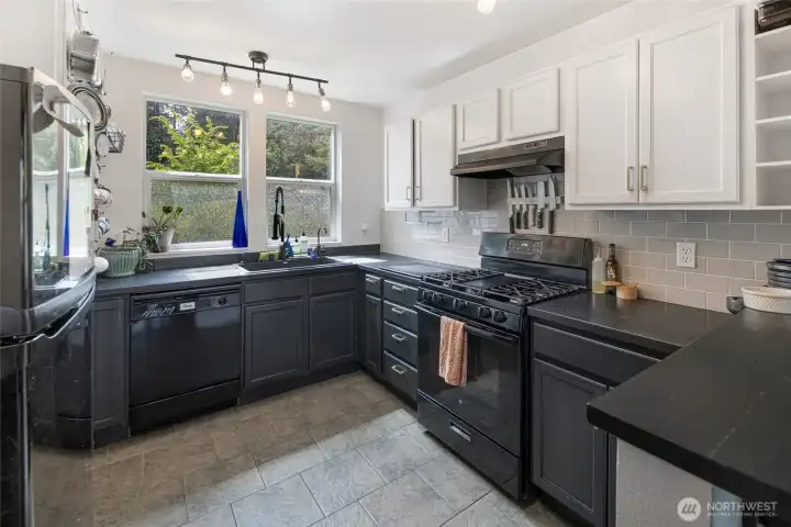 The thoughtfully curated kitchen pairs crisp white upper cabinetry with bold dark countertops & lower cabinets, grey subway tile, and a full suite of black appliances, all bathed in warm natural light from the garden-facing windows.