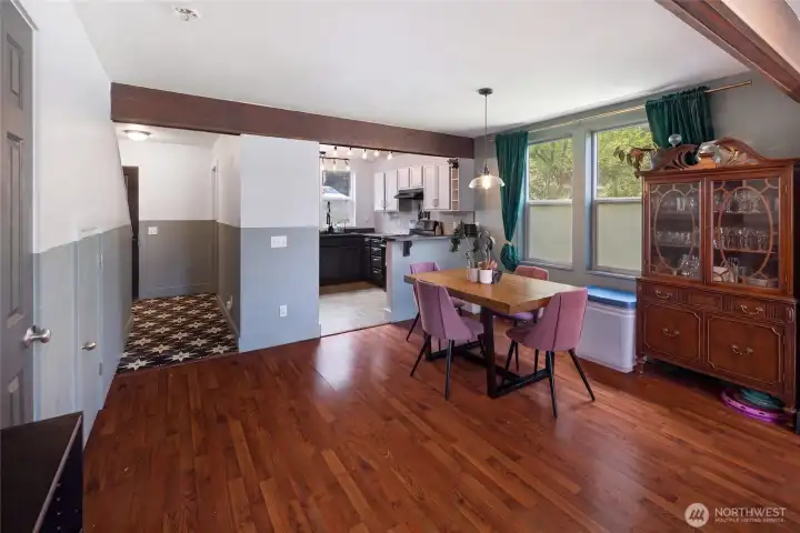The dining area opens directly to the kitchen via a handsome wood beam pass-through, showcasing gleaming laminate floors, two-tone painted walls, and abundant natural light from dual windows. The semi-open floor plan offers seamless flow between living and cooking spaces, with tile flooring in the kitchen and warm wood beam accents tying the areas together.