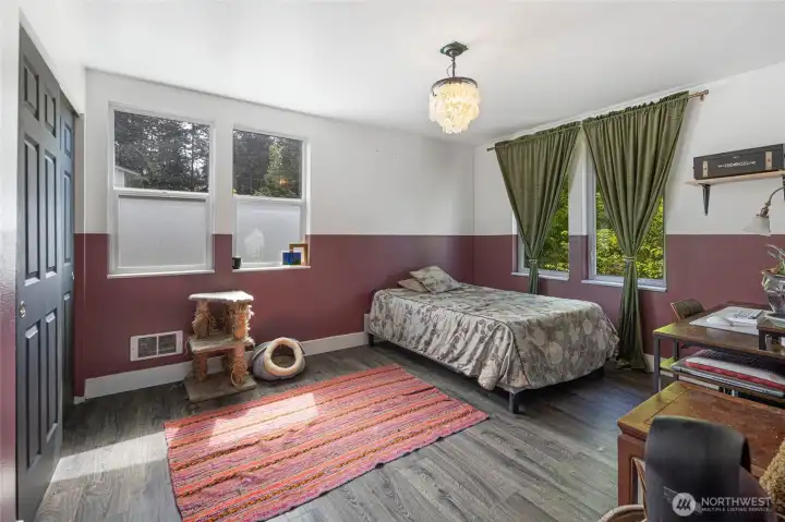 Bathed in natural light from three windows framing lush green treetops, this spacious bedroom feels bright and airy throughout the day — a versatile retreat that effortlessly accommodates a restful sleeping space, creative studio, or elegant home office, all enhanced by a charming capiz shell pendant light and serene, wooded surroundings.