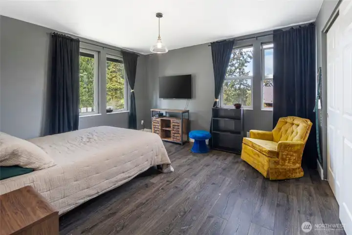 The spacious primary bedroom is bathed in natural light from multiple windows framing lush evergreen views. A cozy yet curated retreat with genuine island character.