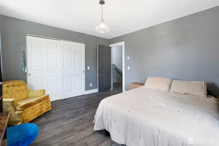 This generously sized primary bedroom features calming grey walls, dark laminate flooring, and a large sliding-door closet offering ample storage for every need. A stylish pendant light anchors the space while the well-proportioned layout and easy hallway access create a functional and inviting retreat.