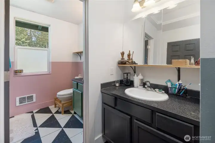 This thoughtfully designed full bath features a split layout with a dedicated vanity area outside the main bathroom. Complete with generous cabinet storage that flows into a whimsical inner bathroom boasting bold black-and-white geometric tile flooring, dusty-rose accent walls, and a green vanity bathed in natural light, offering both privacy and a distinctly charming island aesthetic.